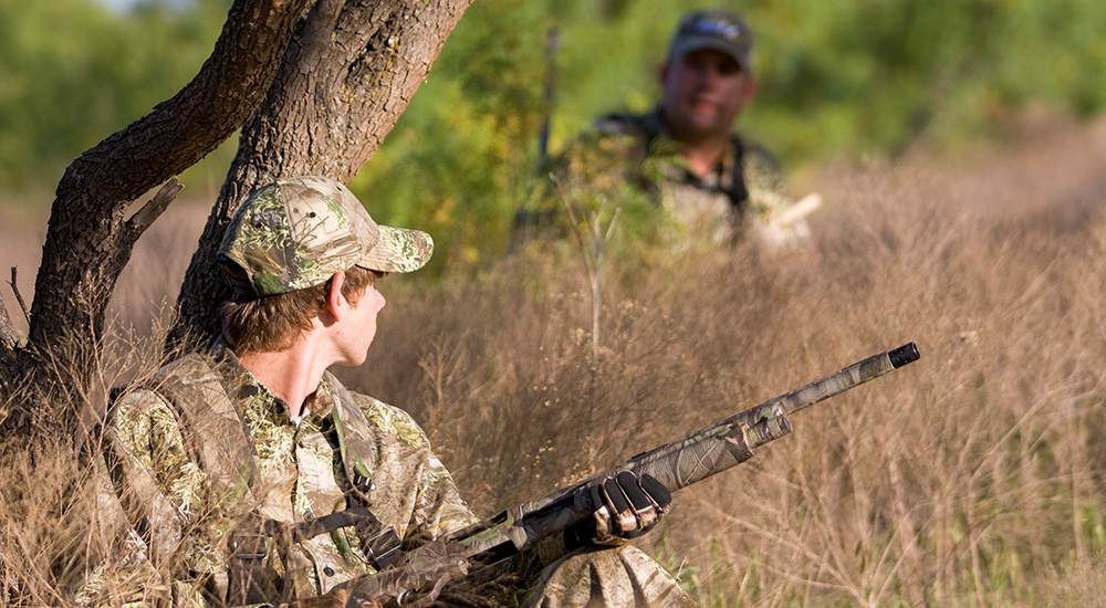 Two male hunters leaning against a tree on a field edge.