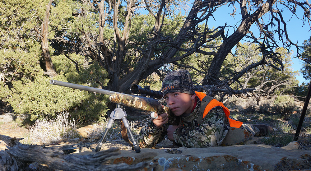 Male hunter in prone position on mountain top preparing to shoot rifle.