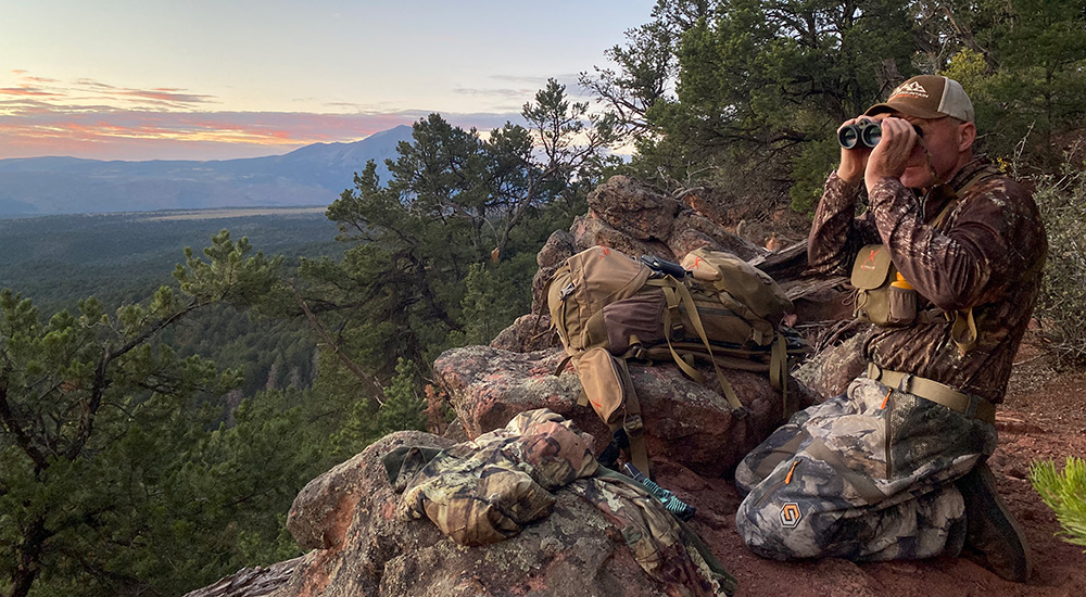 Male hunter using binoculars to spot game at mountain top.