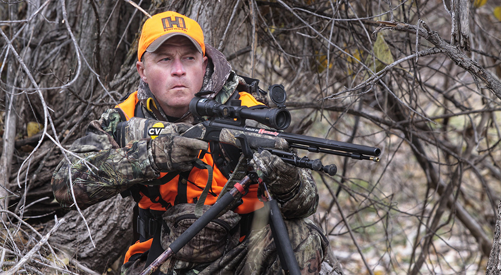 Male hunter holding muzzleloader up to shoulder to prepare for a shot while deer hunting in woods.