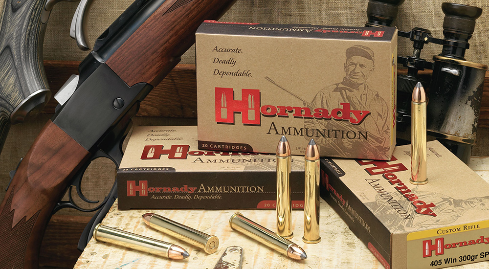 Hornady .405 Winchester ammunition standing on ammunition box with lever-action rifle resting against table.