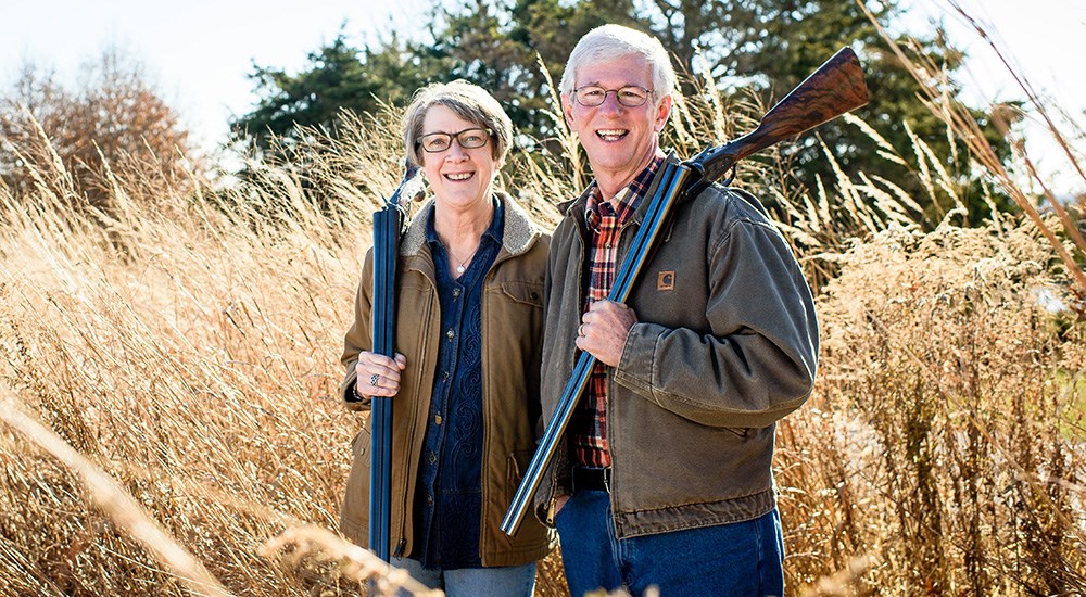 Male and female standing in wheat field with shotguns on their shoulders.