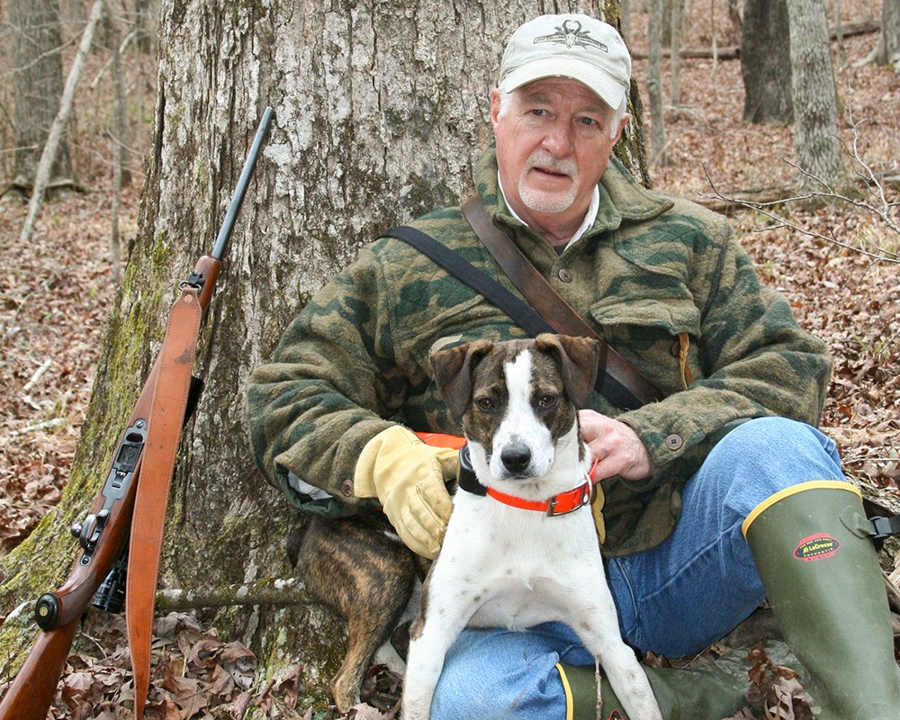 Male hunter with squirrel dog sitting against tree.