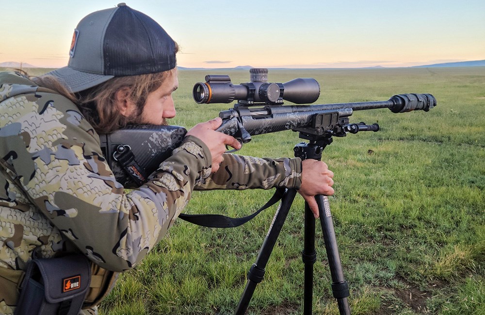 Male hunter shooting Gunwerks suppressed bolt action rifle in open grassy field.