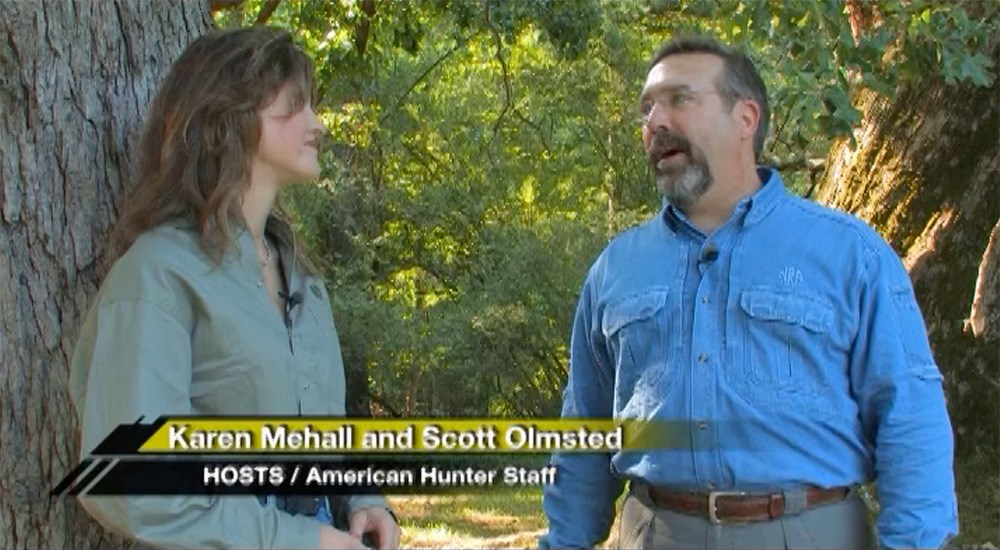 Male and female American Hunter TV hosts talking outdoors next to large tree.