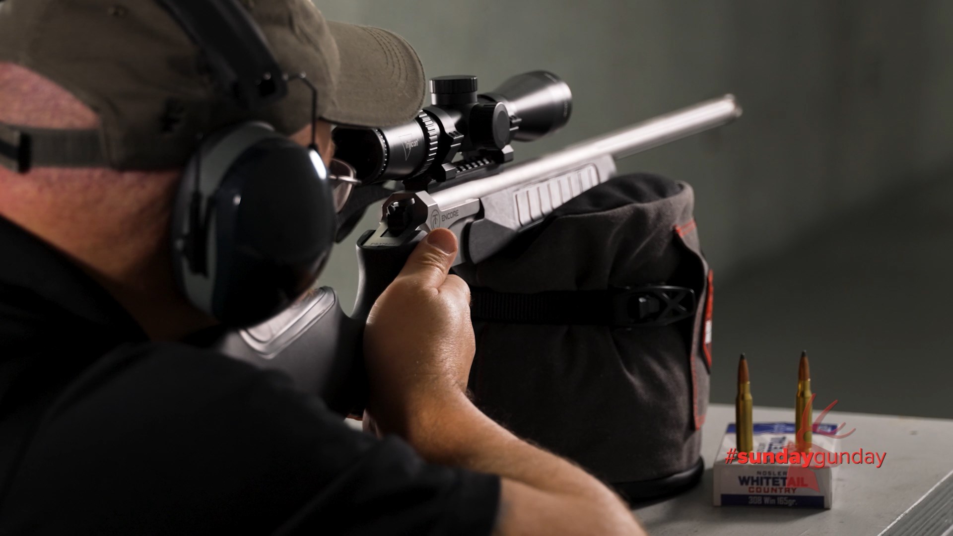 Man wearing protective gear ballcap shooting Thompson/Center Encore single-shot rifle shown with Nosler Whitetail Country packaging and two upright bullets cartridges on indoor dark shooting range with lighting highlights