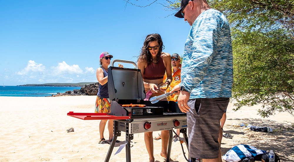Group of friends, male and female, on beach cooking on grill on Molokai Hawaii island.
