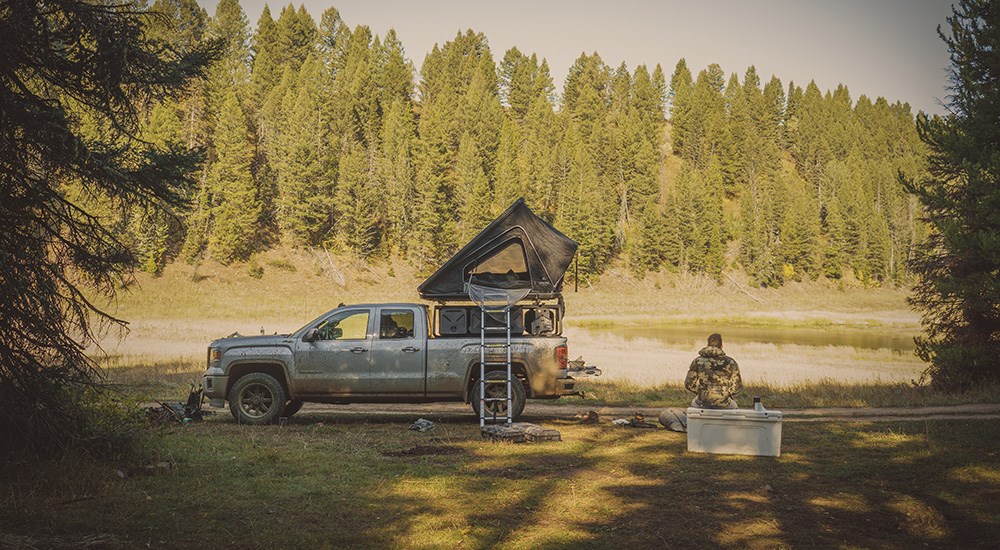 Hunter sitting on cooler beside truck campsite near pond.