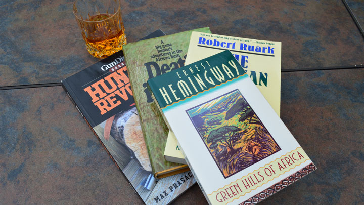 A stack of books with Hemingway&#x27;s &quot;Green Hills of Africa&quot; on top, and a glass of bourbon in the background.