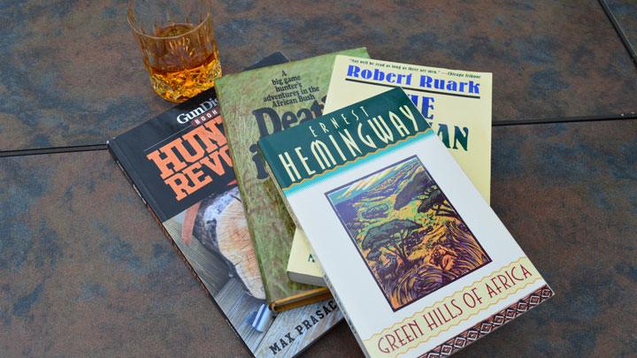 A stack of books with Hemingway's "Green Hills of Africa" on top, and a glass of bourbon in the background.