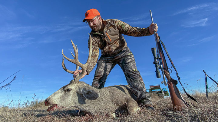 Hunter inspects his quarry: a mule deer on the open plains. A rifle props against the ground in his left hand.