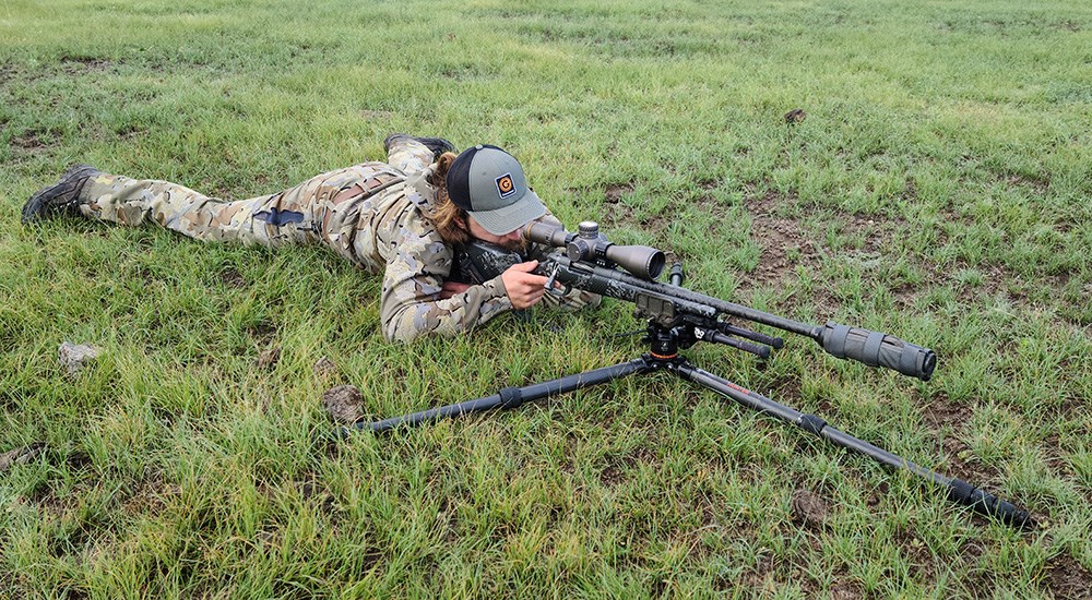 Male hunter shooting bolt action rifle on grass in prone position.
