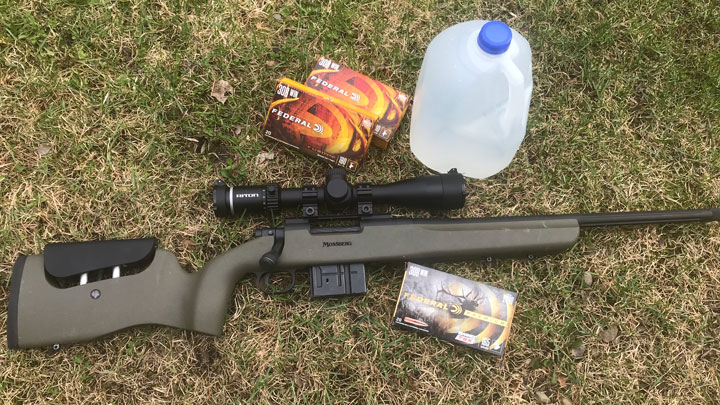 Rifle with optic, ammunition and a water jug all lying in the grass.