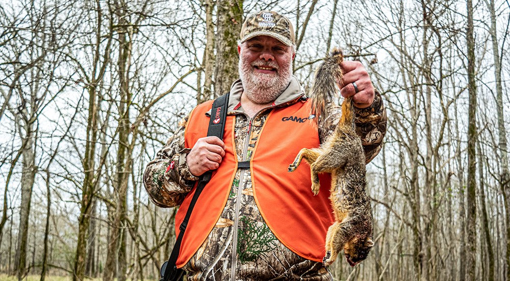 Male hunter wearing camouflage and orange vest holding up squirrel by the tail.