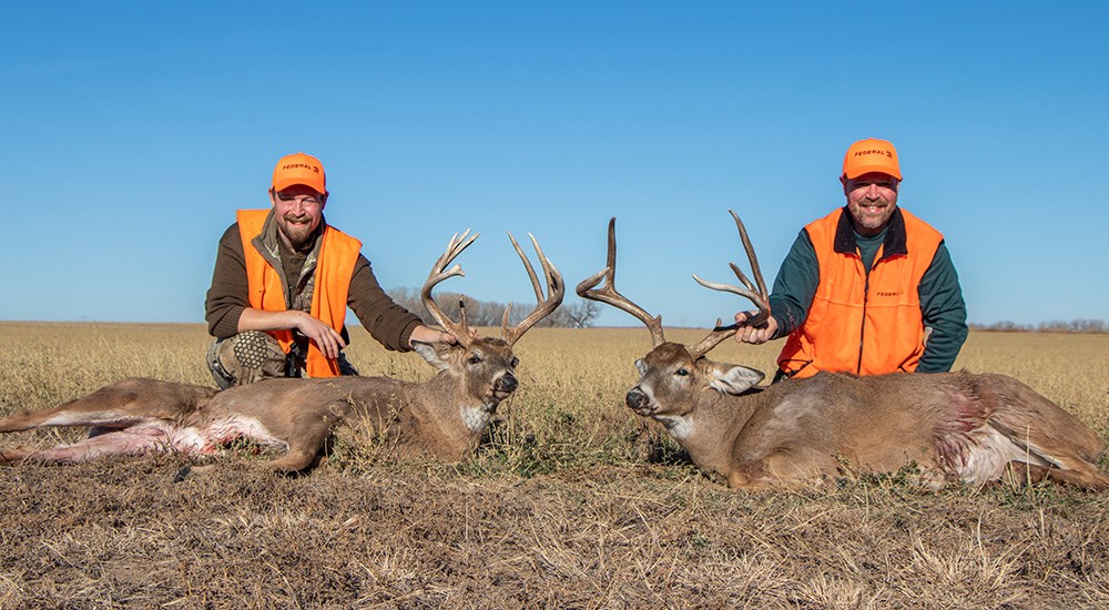 Two male hunters posing with two whitetail bucks in open field.