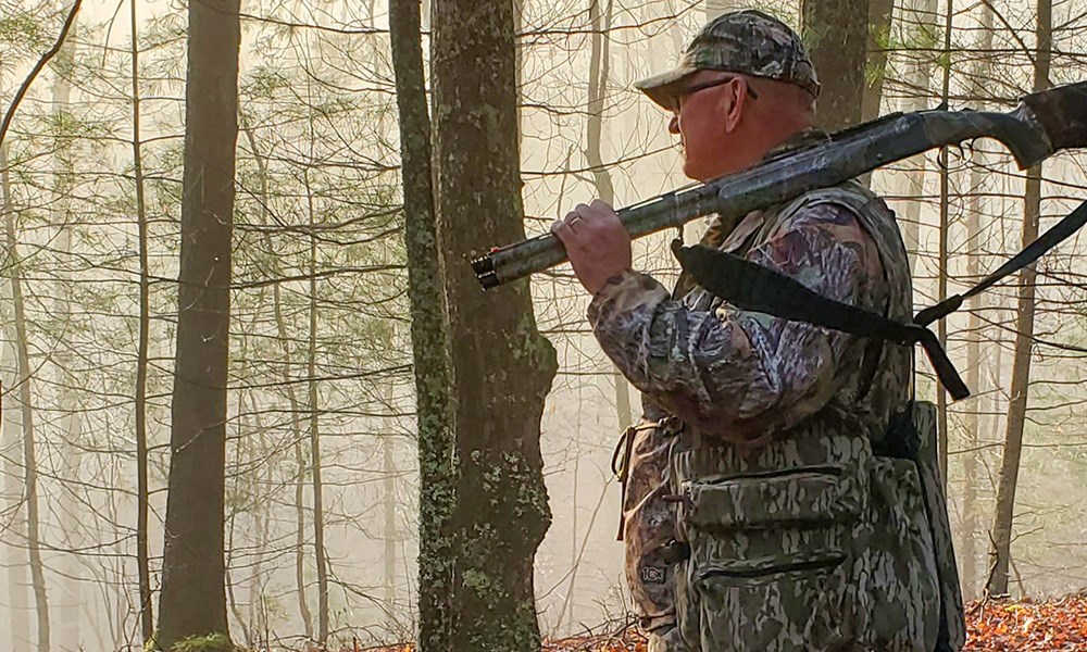 Male turkey hunter in woods with shotgun over shoulder.
