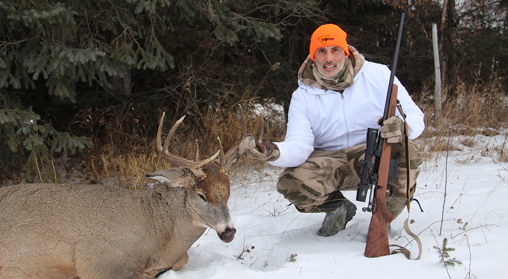 Hunter holding rifle posing with whitetail buck in snow.