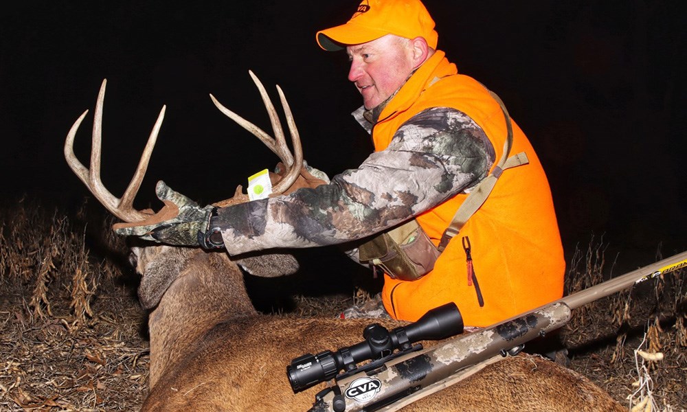 Male hunter in camouflage and orange vest holding whitetail buck.