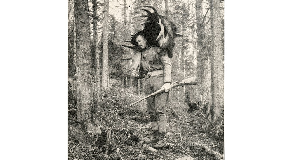 Col. Townsend Whelen in woods with moose antlers on back carrying rifle.