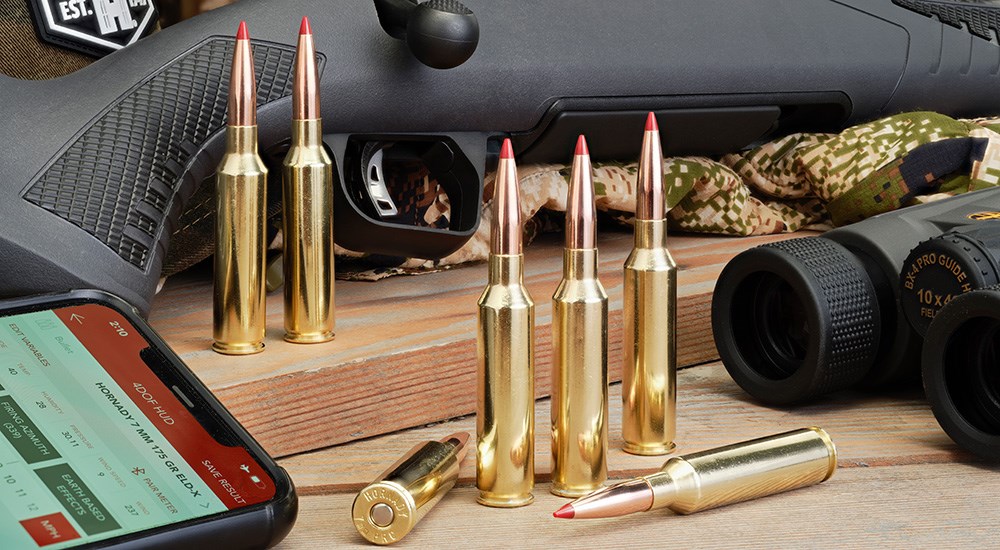 Hornady 7mm Precision Rifle Cartridge ammunition standing up on wood table with Savage bolt-action rifle and Leupold binoculars on right.
