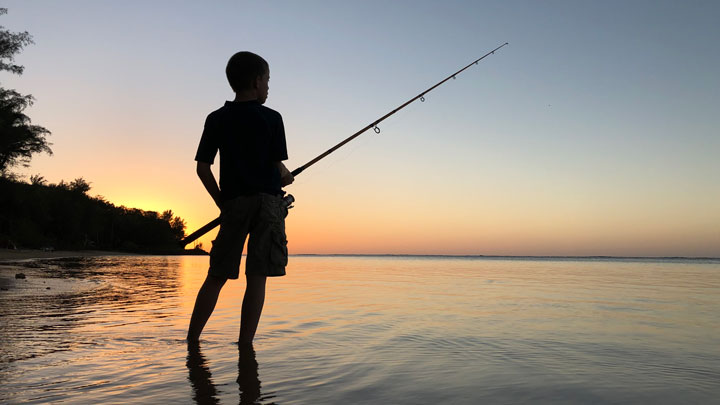 Boy with a fishing rod on a Hawaiian beach stands silhouetted against the setting sun.