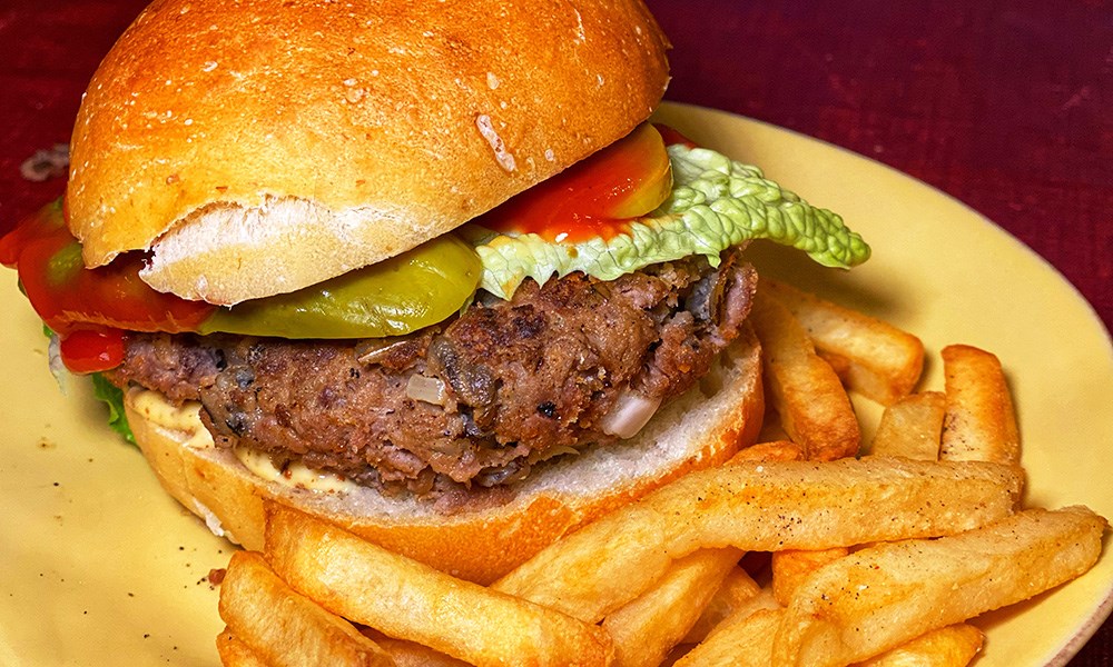 Elk burger topped with lettuce, pickles, and ketchup with French fries on yellow dinner plate.