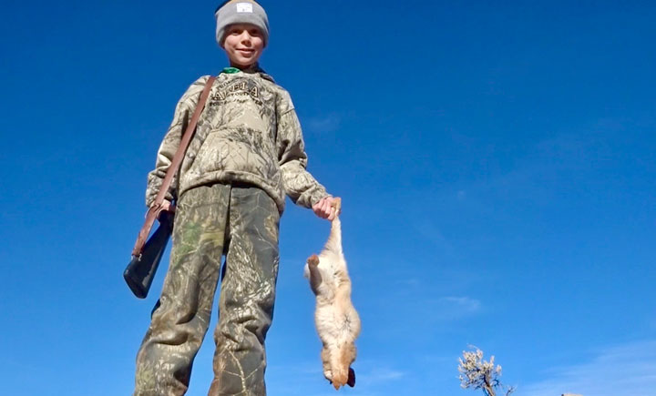 Hunter holding a dead rabiit with a rifle slung over his shoulder, backed by a cloudless deep-blue sky.