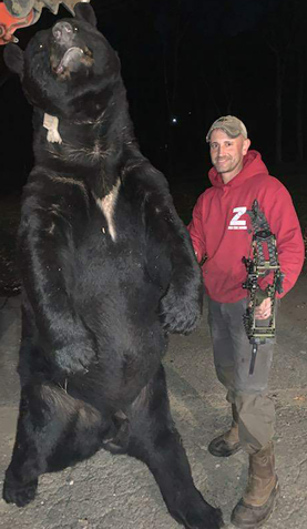 World Record Black Bear, suspended aloft by an orange Kubota, with Jeff standing next to it, bow in hand, in a red sweatshirt
