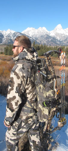 Bowhunter utilizing Bow Spider off his backpack, with snow-capped Western mountains in the background. He stands by a small creek.