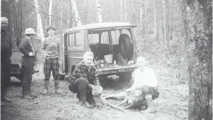 Two Vermont bucks taken, in a black and white photograph with men standing around, and a jeep in the background. The whole scene takes place in the woods.
