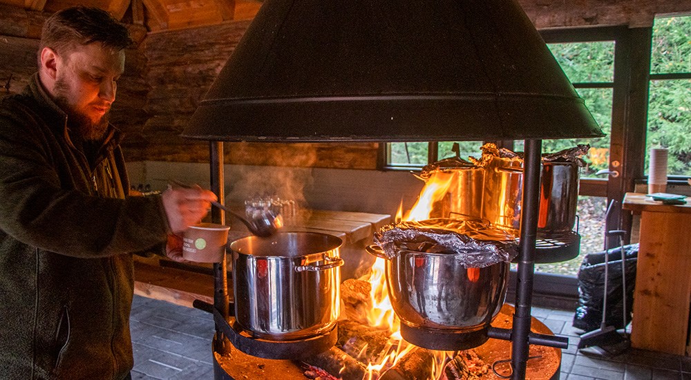 Male using spoon to retrieve soup from stainless steel pot over wood burning fire.