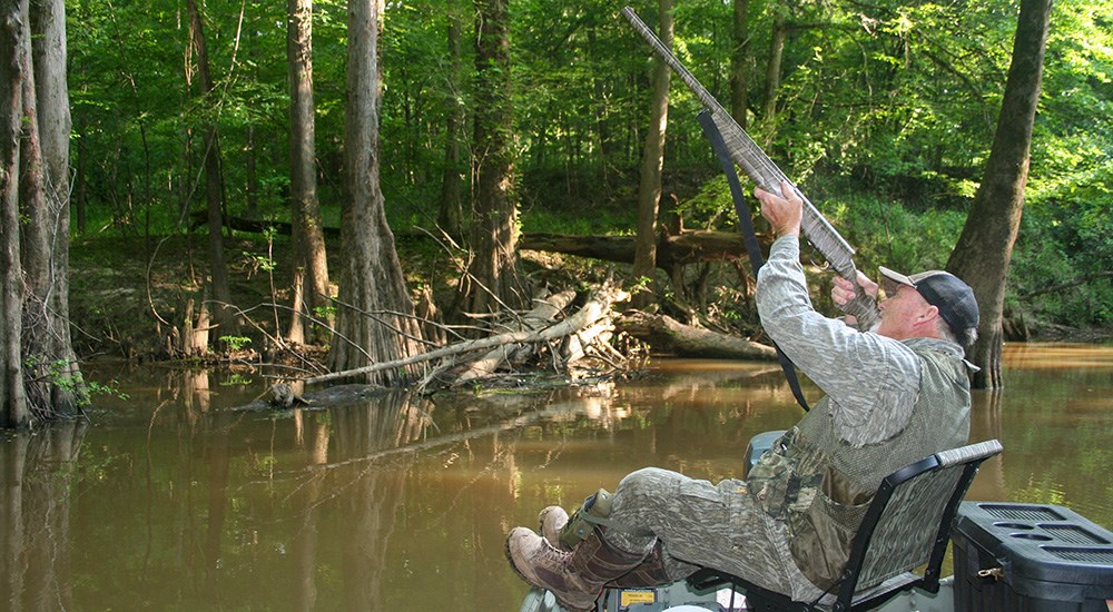 Male hunter in boat on pond shouldering shotgun.