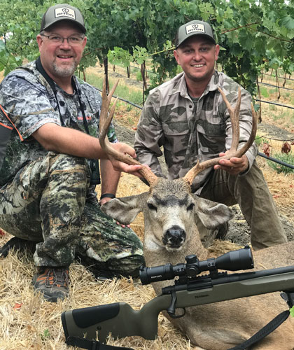 two hunters pose with a downed blacktail, rifle sitting in the foreground.