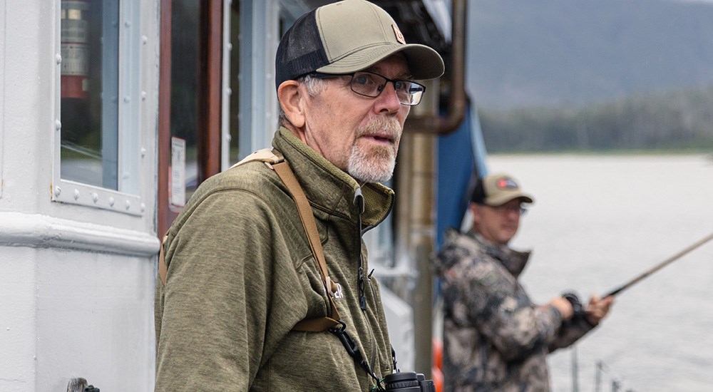 Male in dark green hat and sweatshirt standing on boat deck in Alaska.