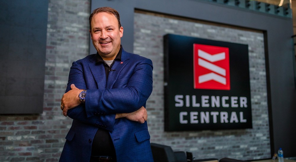 Adult male wearing blue jacket standing with arms crossed in front of Silencer Central logo on brick wall.