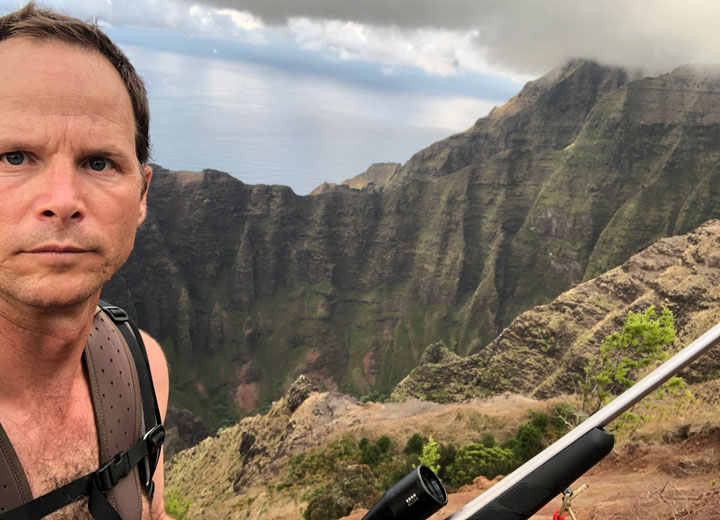 Hunter with pack and rifle looks into camera, with cliffs in the background.