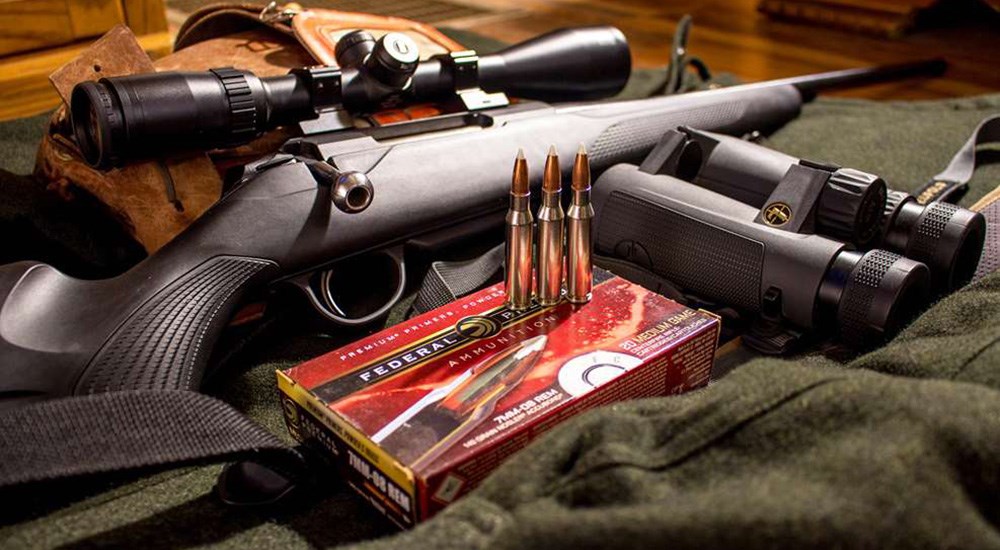 Bolt action rifle with Federal Premium ammunition and Leupold binocular laying on hunter green jacket.