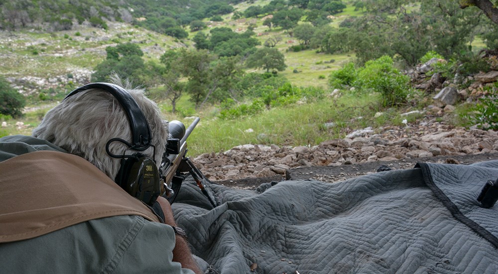 Male with gray hair laying prone shooting rifle at distant outdoor shooting target.