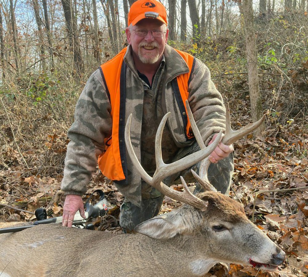 Male hunter wearing camouflage and orange vest and hat posing with whitetail buck.