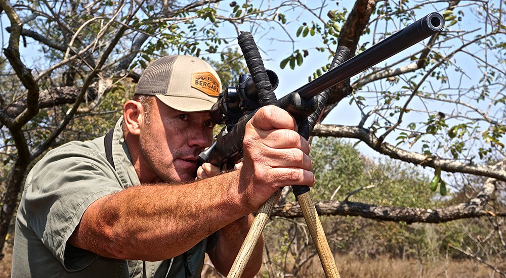 Man standing and shooting rifle off of African shooting sticks