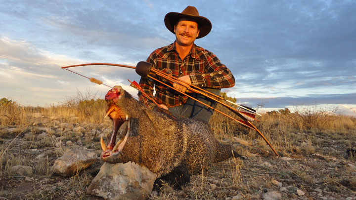 Hunter poses with his handbuilt primitive osage self-bow, behind a massive javie. The javies mouth is open, exposing its cutters, and the sun is setting in the background against the green and brown mesa.
