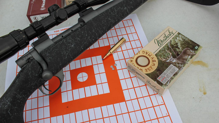 Box of 257 Weatherby and empty shell laying next to grouping shot on target and black bolt-action rifle mounted with scope.