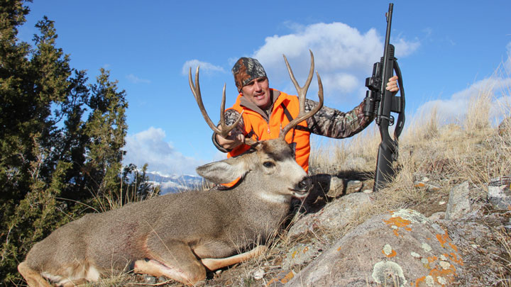 Hunter in orange vest holding buck by antlers. Downed with .308 Winchester rifle in his left hand