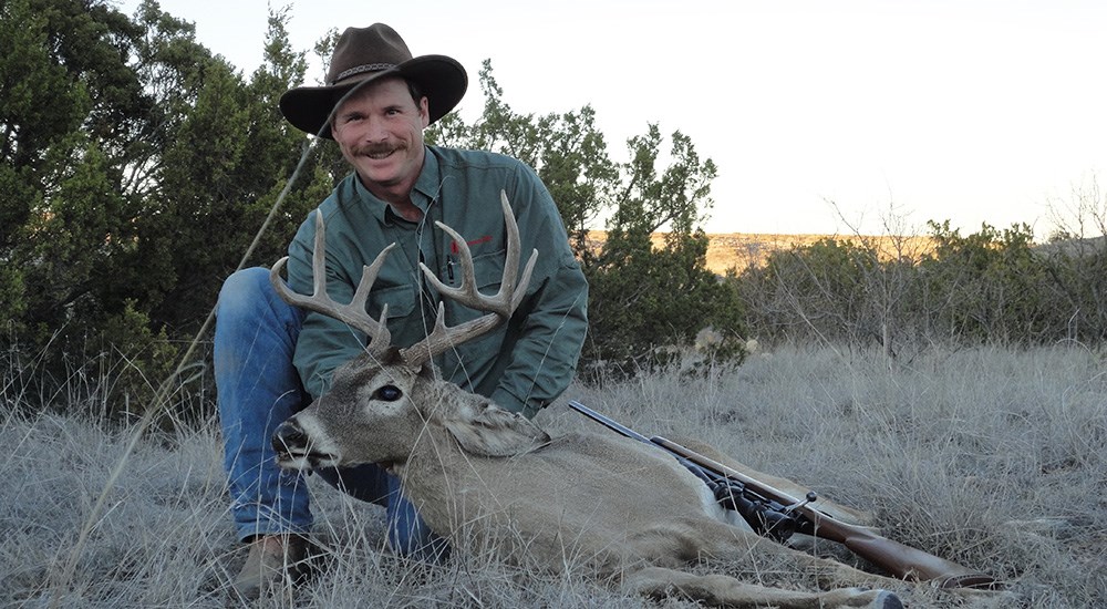 Male hunter wearing blue jeans and dark green shirt posing with whitetail buck.