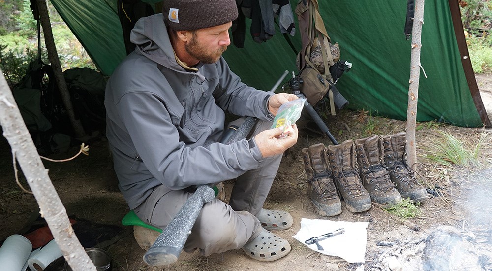 Male sitting inside tarp tent cleaning bolt action hunting rifle.