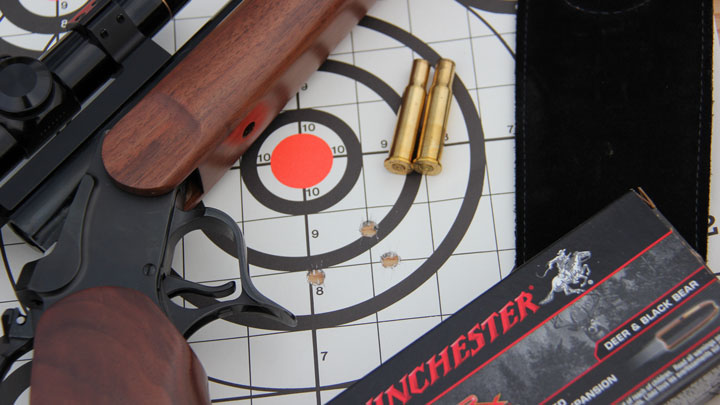 Two .30-30 Winchester shells sitting on a target next to grouping, break-action rifle and ammo box