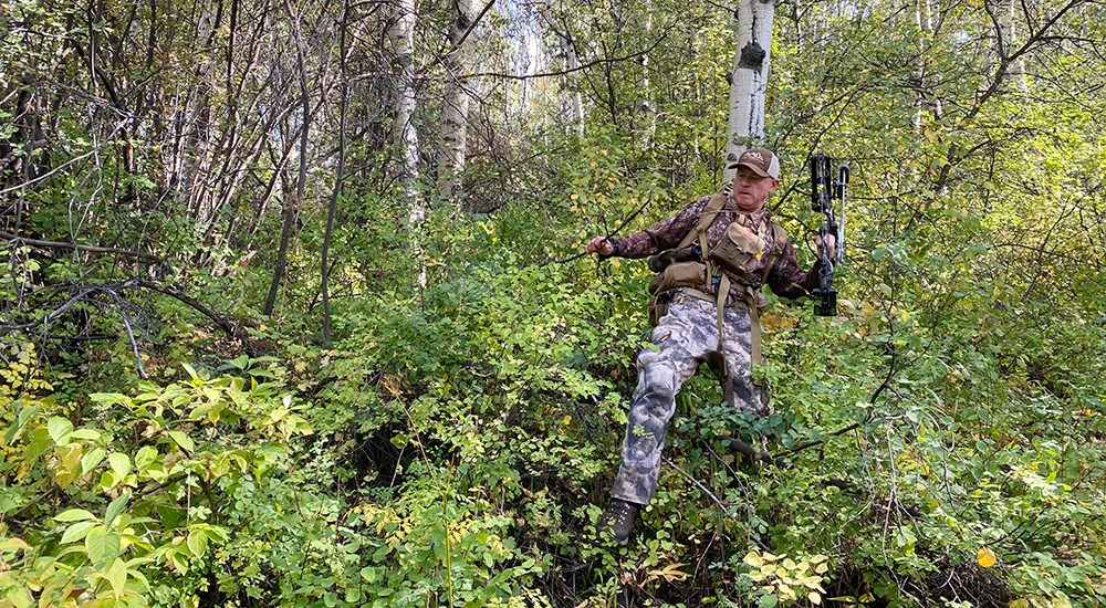 Male hunter carrying compound bow through timber terrain.