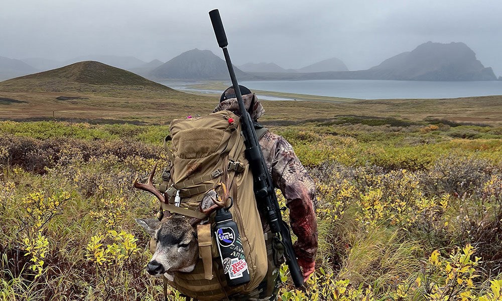 Hunter packing mule deer out of backcountry with suppressed rifle slung on shoulder.