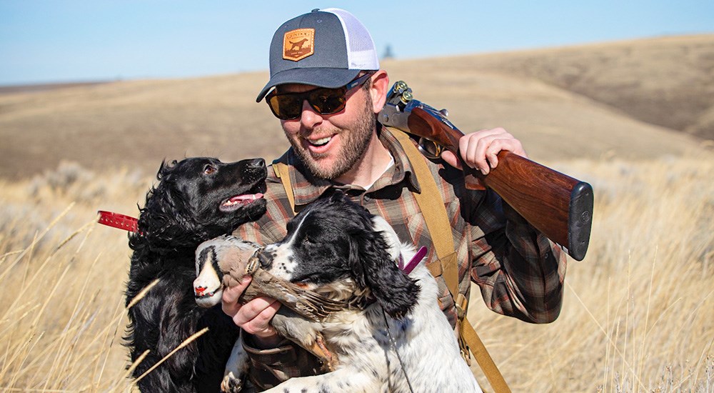 Male wearing plaid shirt with Browning shotgun over shoulder holding chukar with Brittany dog.