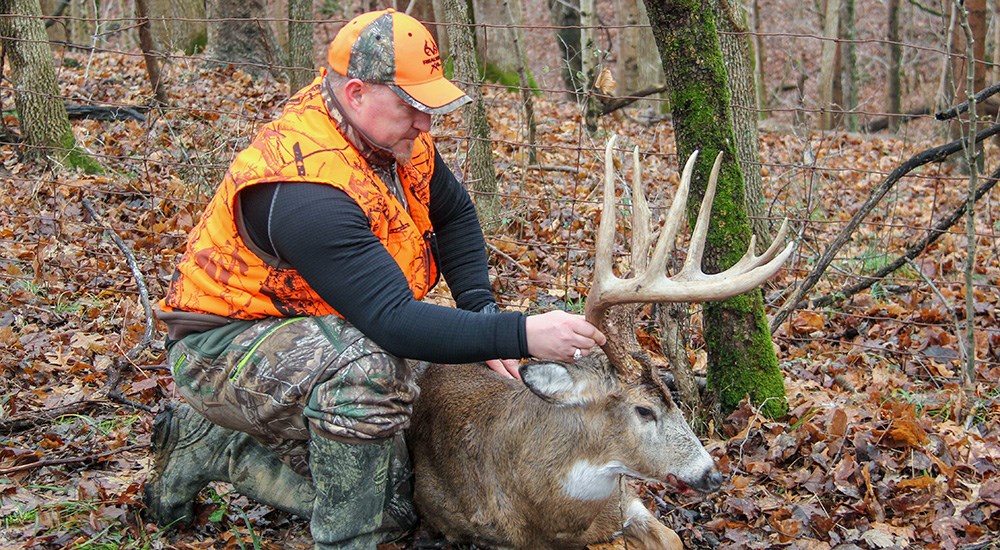 Male hunter wearing camouflage and blaze orange bending down to admire whitetail buck.